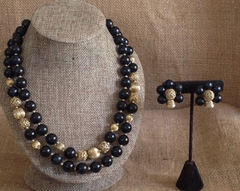 May include: A black and gold beaded necklace and matching earrings. The necklace is double-stranded and features large black beads with gold accents. The earrings are clip-on and feature a similar design.