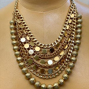 Può includere: Collana dorata a più fili con vari stili e trame di catena. La collana presenta più strati di catene, tra cui catene a maglia, accenti a disco e perline a sfera strutturate. La collana è esposta su un manichino.