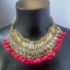 May include: A gold-toned statement necklace with clear rhinestones and red beads. The necklace features a chain, vertical gold bars, and a fringe of red beads. The necklace is displayed on a black mannequin.