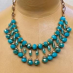 May include: A turquoise beaded necklace with a gold-tone chain. The necklace features multiple strands of faceted turquoise beads and dangling teardrop-shaped beads. Small, clear rhinestones accent the design. The necklace is displayed on a mannequin.