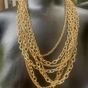 May include: A multi-strand gold-tone necklace displayed on a black mannequin. The necklace features several layers of textured chain links, creating a layered and ornate design. The chains vary in thickness and length, adding visual interest.