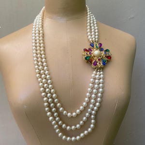 May include: A multi-strand necklace featuring three rows of white pearls. The strands are connected by a large, ornate gold-tone brooch centerpiece adorned with a central white pearl surrounded by faceted red, blue, green, and purple gemstones.