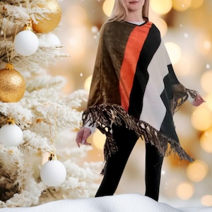 May include: A brown, orange, black, and white striped poncho with fringe trim. The poncho is worn by a person standing in front of a snowy Christmas tree.