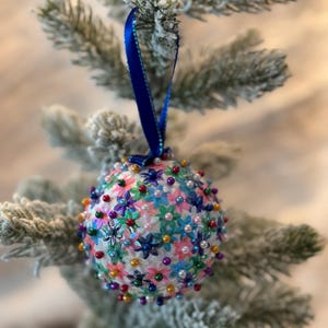 May include: A decorative Christmas ornament, spherical in shape, is covered in small, colorful flowers and pearl-like beads. A dark blue ribbon is attached to the top for hanging. The ornament is set against a backdrop of a snow-covered evergreen branch.