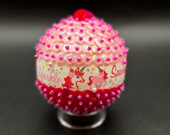 May include: Pink and white Christmas ornament with a unicorn design and the word "Sparkle" on it. The ornament is covered in sequins and has a pink ribbon.