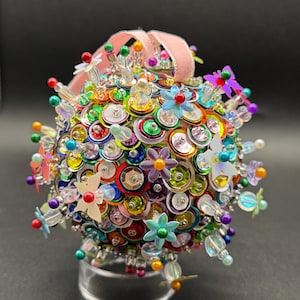 Peut inclure: Une boule ronde colorée recouverte de paillettes, de perles et de petites fleurs. La boule est sur un support transparent et a un ruban rose attaché au sommet.