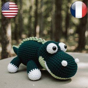 Peut inclure: Un jouet en forme d'alligator crocheté à la main, de couleurs vert foncé et vert clair, avec des détails blancs sur les yeux, les dents et les pieds. L'alligator a une expression amicale et est posé sur un fond naturel flou. Un jouet doux pour enfants.
