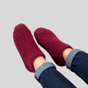 Peut inclure: Une paire de chaussons en laine feutrée bordeaux. Les chaussons sont portés avec un jean bleu dont les revers sont retroussés. Le fond est gris clair uni.