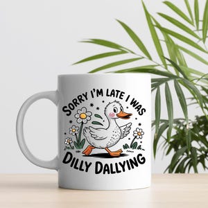 Peut inclure: Une tasse en céramique blanche avec une illustration en noir et blanc d'un canard marchant dans un champ de fleurs. Le texte "Sorry I'm late I was dilly dallying" est imprimé sur la tasse.