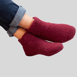 Peut inclure: Une paire de chaussons en laine feutrée bordeaux. Les chaussons sont d'un rouge profond et semblent doux et chauds. La personne porte un jean bleu.