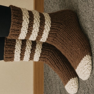 Peut inclure: Une paire de chaussettes en tricot marron avec des rayures blanches en haut et des orteils blancs. Les chaussettes sont épaisses et semblent faites à la main, parfaites pour le froid. Elles sont en matière douce et chaude.