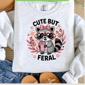 Peut inclure: Un sweat-shirt gris clair avec un raton laveur de dessin animé portant des lunettes de soleil en forme de cœur et un nœud rose. Le raton laveur tient une boisson rose, entouré de feuilles et d'étoiles roses. Le texte "CUTE BUT FERAL" est au-dessus et en dessous du raton laveur.