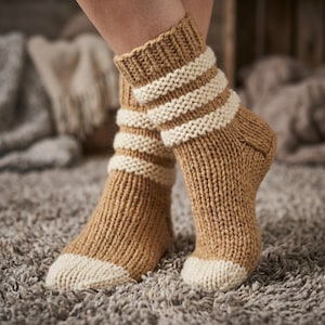 May include: A pair of knitted socks in a warm tan color with cream-colored stripes and toe caps. The socks are thick and appear cozy, perfect for cold weather. The ribbed texture adds to their handmade appeal.