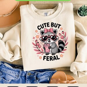 Peut inclure: Sweat-shirt crème avec un dessin de raton laveur portant des lunettes de soleil en forme de cœur et tenant une boisson. Le texte "CUTE BUT FERAL" est imprimé en dessous. L'image comprend également le texte "INSTANT DOWNLOAD" et des icônes de type de fichier.