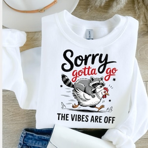 Peut inclure: Sweat-shirt blanc avec un raton laveur noir et gris chevauchant un poulet blanc. Le texte "Sorry gotta go * THE VIBES ARE OFF" est imprimé sur le sweat-shirt.