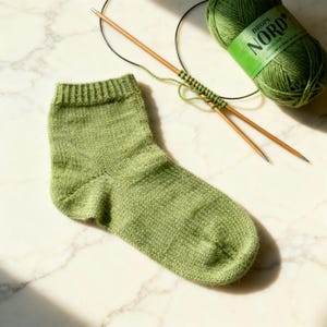 Peut inclure: Une chaussette verte tricotée terminée avec des aiguilles à tricoter en bois et une pelote de laine verte. L'étiquette de la laine indique "DROPS NORD". La chaussette est sur une surface en marbre blanc.