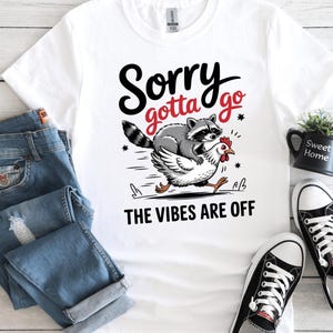 Peut inclure: T-shirt blanc avec un dessin d'un raton laveur chevauchant une poule, avec le texte "Sorry gotta go" et "The Vibes Are Off". L'image comprend un jean bleu et des baskets noires.