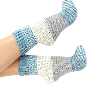 Peut inclure: Une paire de chaussettes en crochet bleues, blanches et grises. Les chaussettes présentent un motif texturé avec un revers bleu, un centre blanc et un talon et un bout gris. Les chaussettes sont présentées sur fond blanc.