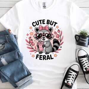 Peut inclure: T-shirt blanc avec un raton laveur de dessin animé portant des lunettes de soleil en forme de cœur et un nœud rose. Le raton laveur tient une boisson rose avec une paille. Le texte "CUTE BUT FERAL" est au-dessus et en dessous du raton laveur.