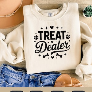 Peut inclure: Sweat-shirt crème avec l'inscription noire "TREAT Dealer", entourée d'empreintes de pattes, de cœurs, d'étoiles et d'os. Le sweat-shirt est posé sur un jean bleu, un chapeau et des sandales.