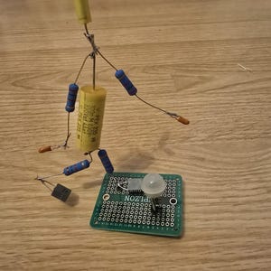 May include: A circuit board with a small LED light, a capacitor, resistors, and a transistor. The circuit board is green and has a grid of holes for soldering components. The components are connected with wires.