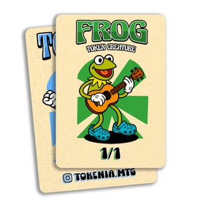 May include: Two trading cards featuring a cartoon frog playing a ukulele. The frog is green with white gloves and blue shoes. The cards have the text "FROG TOKEN CREATURE" and "1/1". The cards are on a beige background.