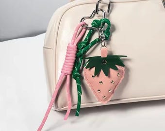 Könnte beinhalten: Ein rosa erdbeerförmiger Schlüsselanhänger mit silbernen Nieten und einem grünen Blattoberteil. Der Schlüsselanhänger ist an einer weißen Handtasche mit einem herzförmigen Silberverschluss und rosa und grünen Seildetails befestigt.