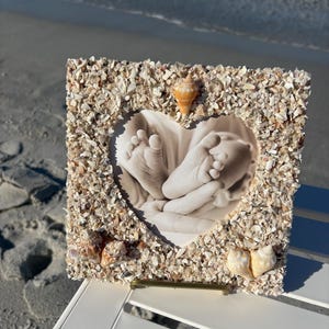 Op de afbeelding: Een vierkante fotolijst versierd met schelpen en kleine schelpfragmenten. De lijst bevat een zwart-witfoto van babyvoetjes in een hartvormige uitsparing. Verschillende schelpen zijn aan de lijst bevestigd.