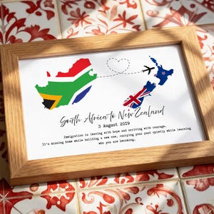 Könnte beinhalten: Ein gerahmter Druck in einem hellen Holzrahmen. Der Druck zeigt eine Karte von Südafrika und Neuseeland, verbunden durch eine gestrichelte Linie und ein Herz, mit dem Text "South Africa to New Zealand" und dem Datum "3. August 2019".