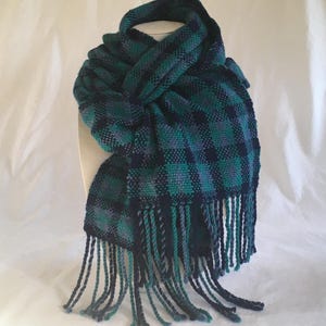Puede incluir: Bufanda tejida a cuadros en tonos verde azulado, azul marino y morado. La bufanda tiene flecos en los extremos. La bufanda se muestra sobre un fondo blanco.