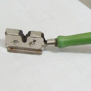 May include: A vintage knife sharpener with a silver metal body and a bright green handle. The sharpener has a double-sided design with two sharpening slots. The handle is rounded and appears to be made of wood or a similar material.