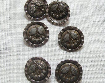 May include: Six antique silver-toned buttons with a detailed botanical design. Each button features a central motif of leaves and berries within a scalloped edge. The buttons are arranged on a white, textured fabric.
