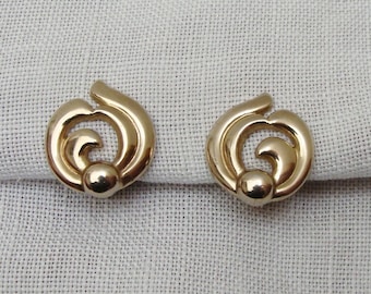 May include: A pair of gold-colored stud earrings. Each earring features a swirling, open design with a small, round accent at the center. The earrings are displayed on a light-colored, textured fabric.