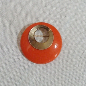 May include: A round, orange brooch with a gold-colored center ring and pin. The brooch has a glossy finish and is set against a white background. The design is simple and geometric.