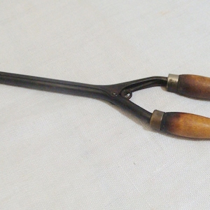 May include: An antique curling iron with a dark metal body and two wooden handles. The handles are a light brown color with a darker, burnt-looking area near the metal. The curling iron is resting on a white surface.