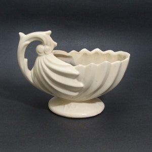 May include: Cream-colored ceramic planter with a seashell design. The planter has a scalloped rim and a decorative handle. It is raised on a small, round base. The overall shape is reminiscent of a clam shell.