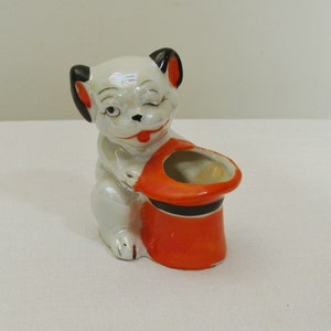 May include: A ceramic figurine of a winking puppy holding an orange and black top hat. The puppy is white with black ears and a red tongue. The hat has a black band.