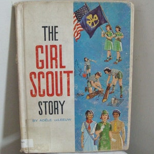May include: Vintage book titled "The Girl Scout Story" by Adèle DeLeeuw. The cover features illustrations of Girl Scouts, an American flag, and a Girl Scout flag. The book has a worn, off-white cover with red and black text.