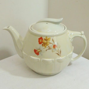 May include: Cream-colored ceramic teapot with a floral design featuring orange, yellow, and white flowers. The teapot has a curved spout, a loop handle, and a lid with a leaf-shaped handle. The base has a scalloped design.