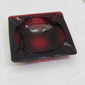 May include: Square, ruby-red ashtray with a rounded interior bowl. The ashtray has a glossy finish and a slightly raised rim. The color transitions from a deep red to a lighter shade in the center. A vintage item.