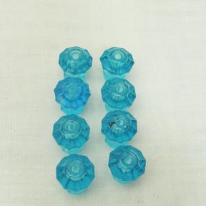 May include: Eight vintage, faceted, turquoise glass cabinet knobs. Each knob has a rounded, octagonal shape with a central indentation. The knobs are arranged in two columns of four on a white background.