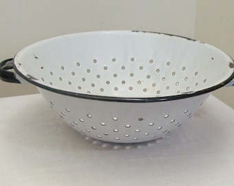 May include: White enamel colander with black trim and handles. The kitchenware item has numerous small holes throughout the bowl for draining liquids. The colander is a classic kitchen tool for washing and preparing food.