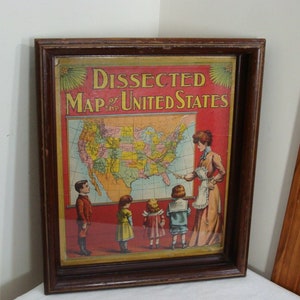 May include: Framed vintage educational poster titled "Dissected Map of the United States." The map shows the U.S. with a teacher and children. The frame is dark brown.