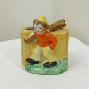 May include: A vintage ceramic figurine of a young boy carrying wood, set against a yellow tree trunk-shaped vase. The boy wears an orange shirt, yellow pants, and a matching hat. The base features green grass and black shoes.