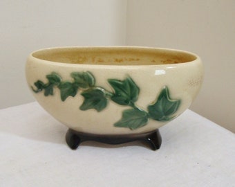 May include: Cream-colored ceramic planter with a green ivy leaf design. The oval-shaped planter sits on three black feet. The interior of the planter is a darker cream color.