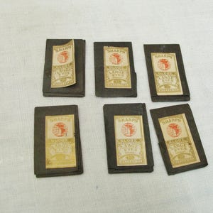 May include: Six vintage packets of Sharps Globe Large Eyes needles. Each packet is dark gray with a cream-colored label featuring a globe graphic and text. The packets are arranged on a white surface.