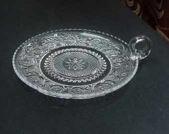 May include: Clear glass oval serving tray with a handle. The tray features an ornate, embossed floral and starburst design. The rim has a scalloped edge. The handle is a small, clear loop. The tray is likely vintage.