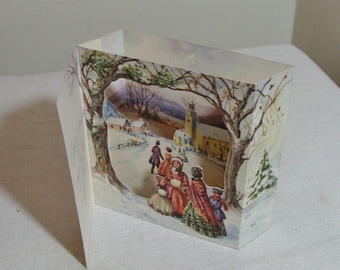 May include: A three-dimensional paper Christmas card depicting a winter scene. The card features a cutout showing a snowy village with figures in period clothing, a church, and trees. The card is open and standing upright.