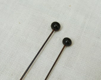May include: Two antique hat pins with black, round heads and long, thin, dark brown stems. The pins are set against a white, textured fabric background. The pins are likely used for securing hats or other accessories.