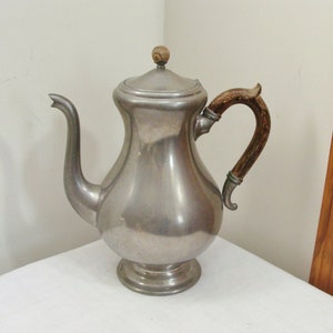 May include: A vintage silver-colored metal coffee pot with a wooden handle and lid. The pot has a bulbous body, a curved spout, and a round base. The handle is made of wood and is attached to the pot with metal fittings. The lid is topped with a small wooden knob.